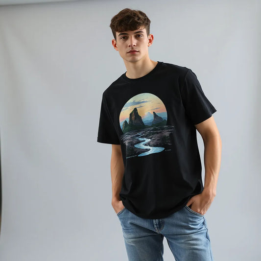 Model in a black surreal graphic tee with a dreamlike melting landscape illustration against a gray studio backdrop